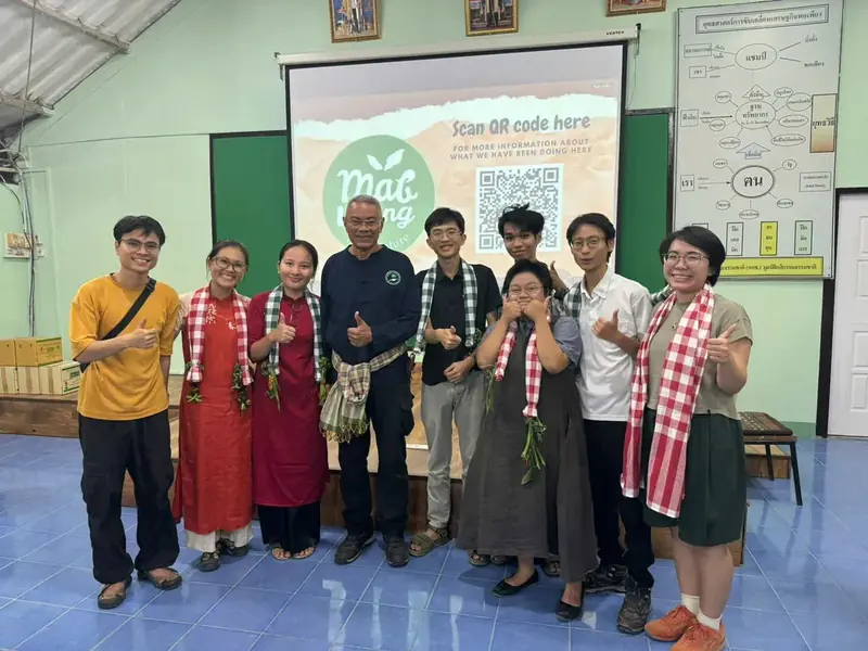 Chín người đứng trong phòng, một số đeo khăn sọc đỏ trắng và giơ ngón cái lên, phía sau là màn hình có mã QR và logo 'mak long'.